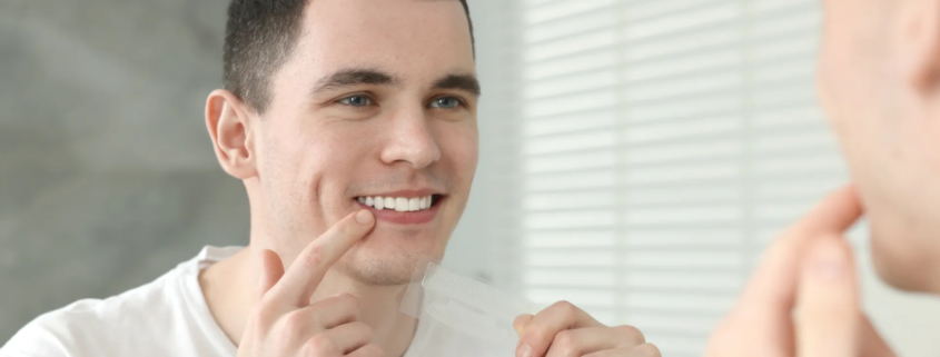 Homme souriant devant un miroir en appliquant une bande de blanchiment dentaire sur ses dents.